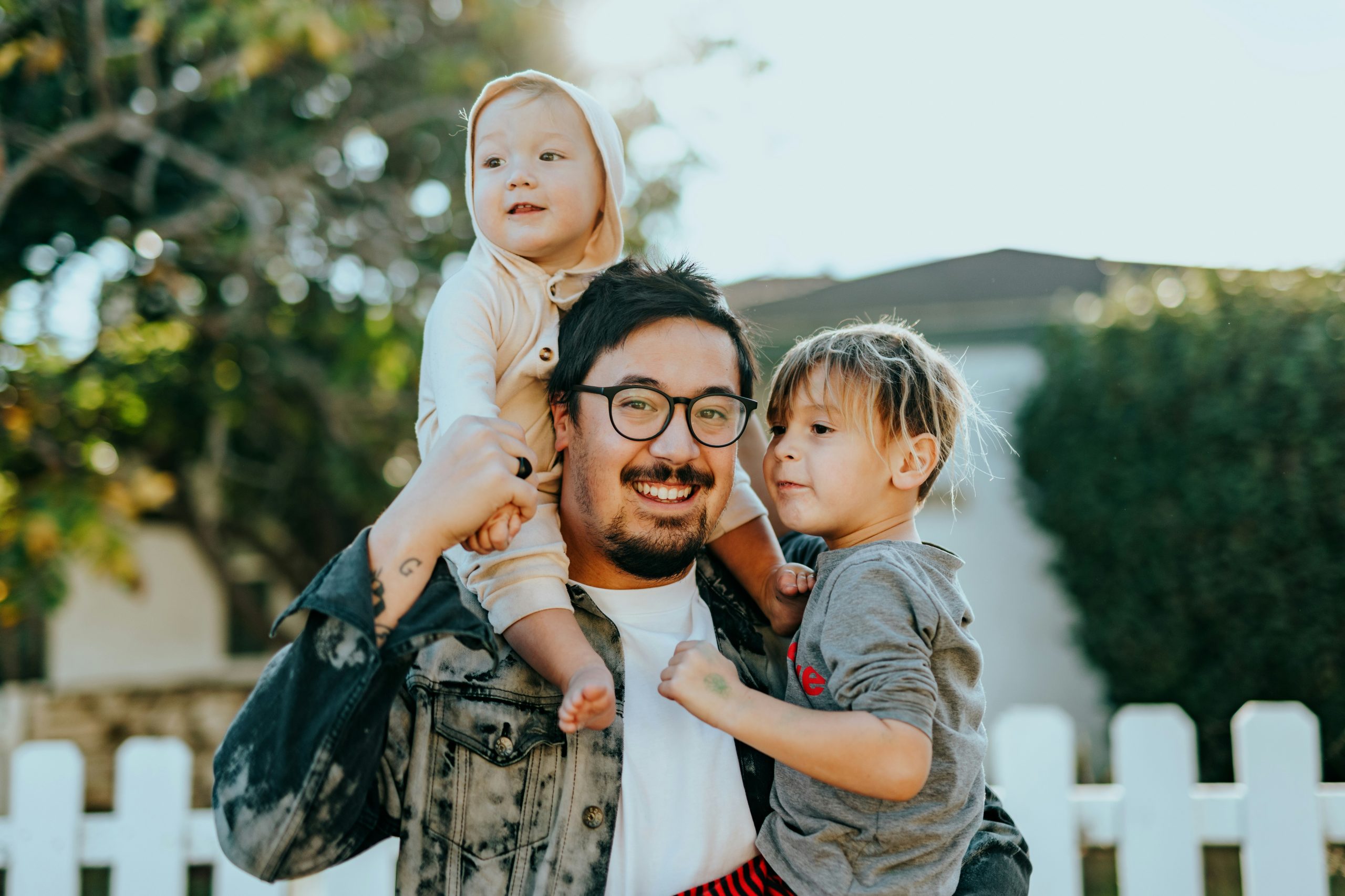 Happy male presenting parent carrying his two children outdoors with one up on his shoulders